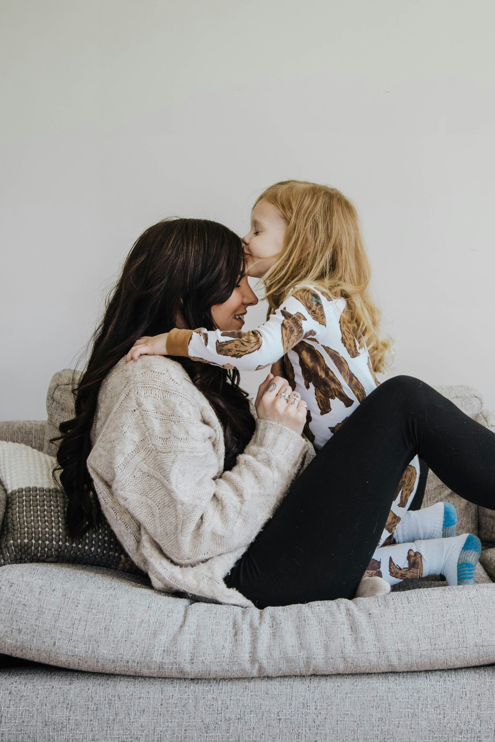 Mom and daughter sharing a cozy moment on the couch, with the daughter laughing and kissing her mom's forehead.