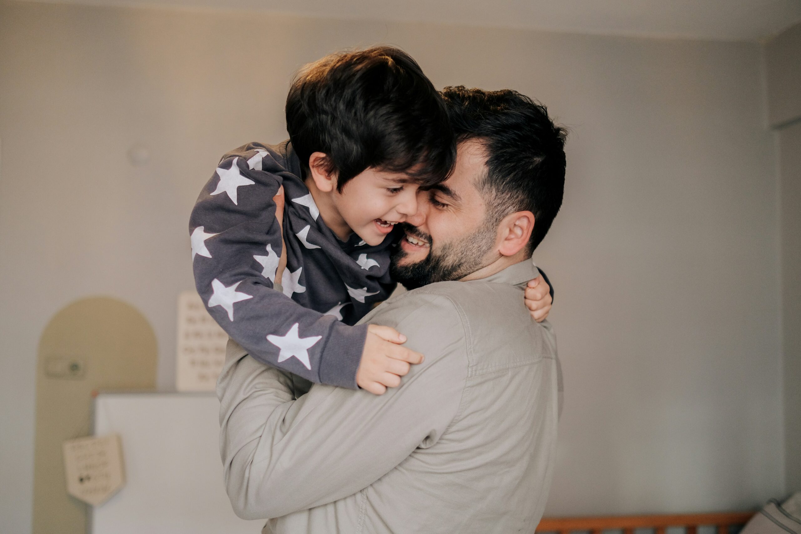 A dad playfully holding his laughing son in his arms while they enjoy a joyful moment together in the son’s room.